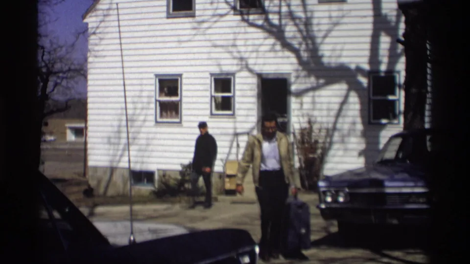 In Madison, Wisconsin, 1966, a man carries a purple suitcase from a white-sided house toward a dark sedan while another man stands nearby.