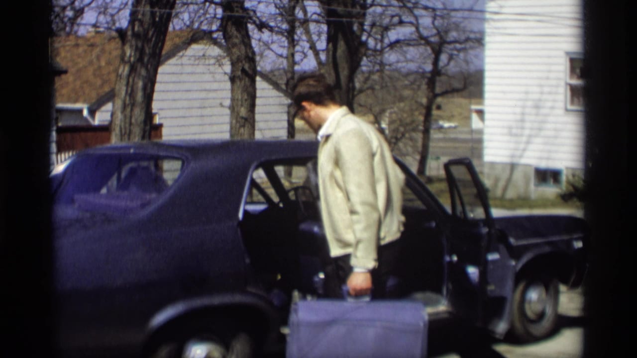 A man in a cream jacket stands by the open rear door of a dark blue sedan in a Madison driveway, suitcase in hand.