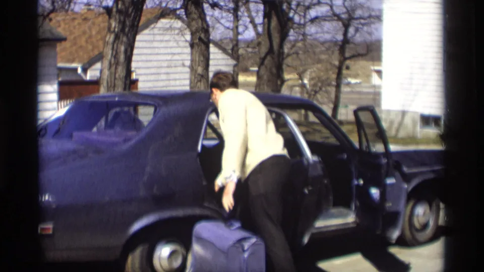 The man bends at the side of the dark sedan, preparing to place the purple suitcase into the rear seat area.