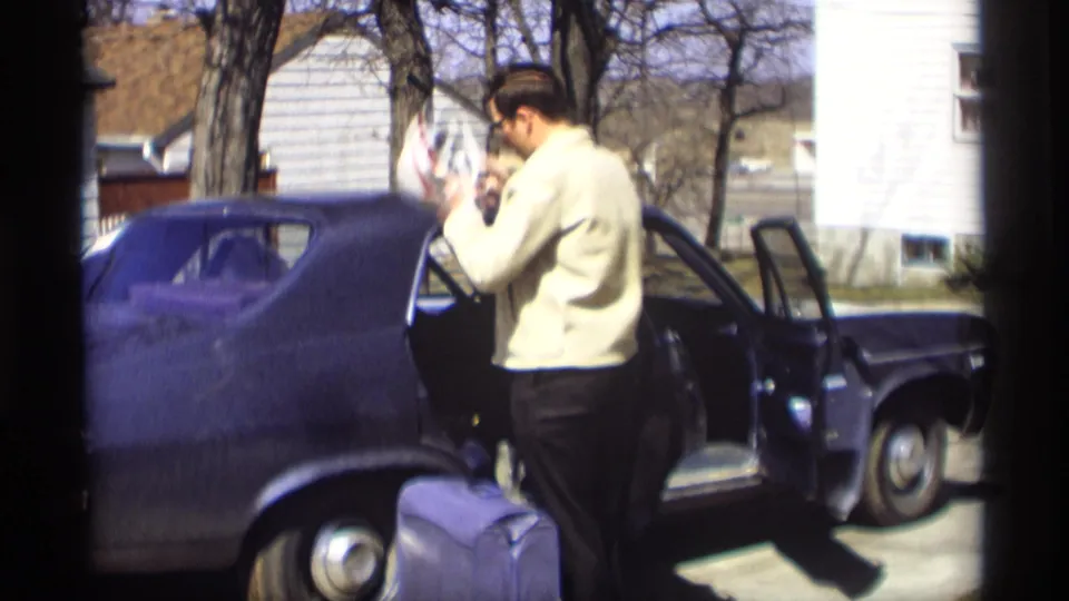 Beside the open rear door of the dark sedan, the man lifts a light patterned item while the suitcase remains on the driveway.