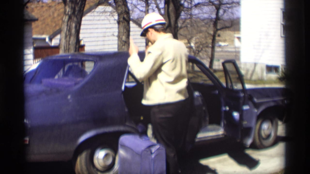 In Madison, Wisconsin, 1966, a man in a light jacket and striped cap stands by an open dark sedan door with a purple suitcase.