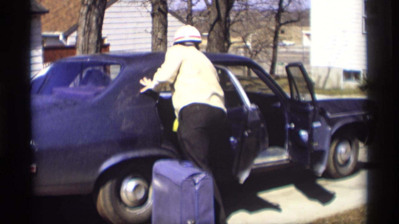 Man leans toward the side of a dark sedan with the driver's door open and suitcase on the ground.