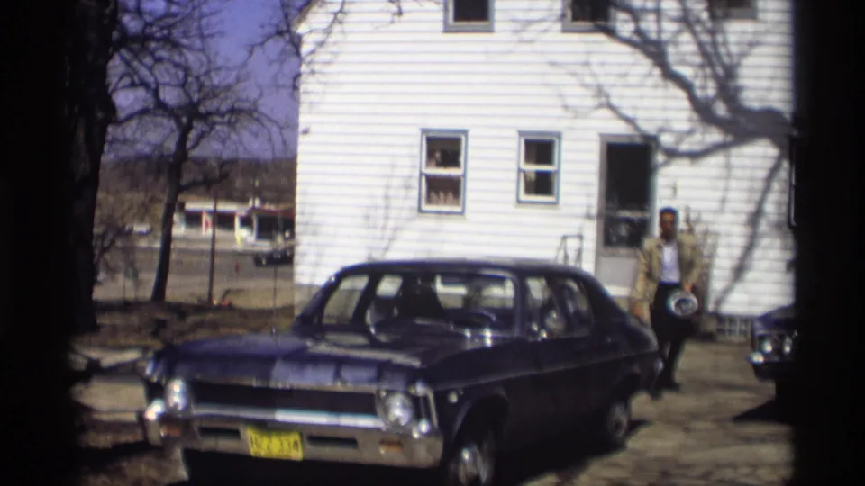 The dark sedan is parked before a white house in Madison, Wisconsin, as the man moves near the entrance during the 1966 home movie.