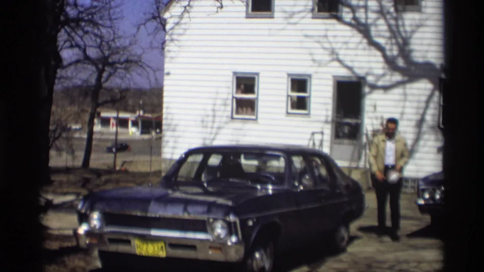 A man stands by the white house behind a parked dark sedan, with driveway, tree shadows, and neighboring homes visible in Madison, 1966.