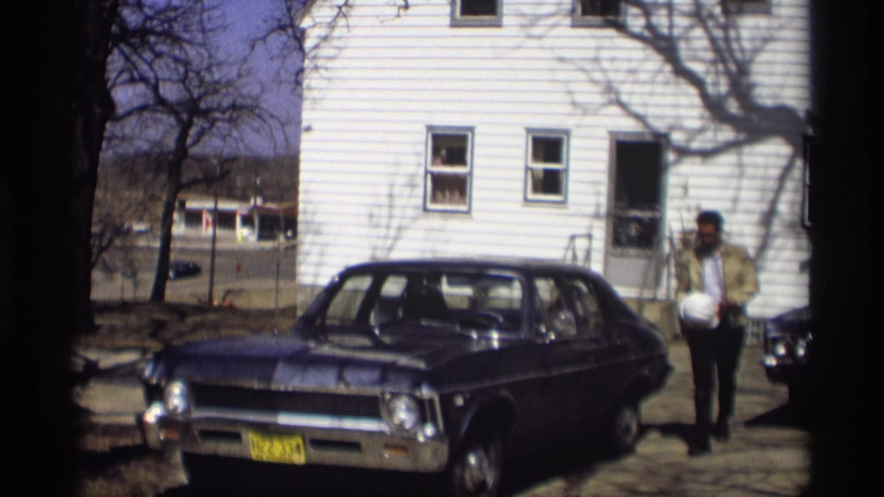 Man in a tan jacket walks beside a blue sedan in front of a white clapboard house.