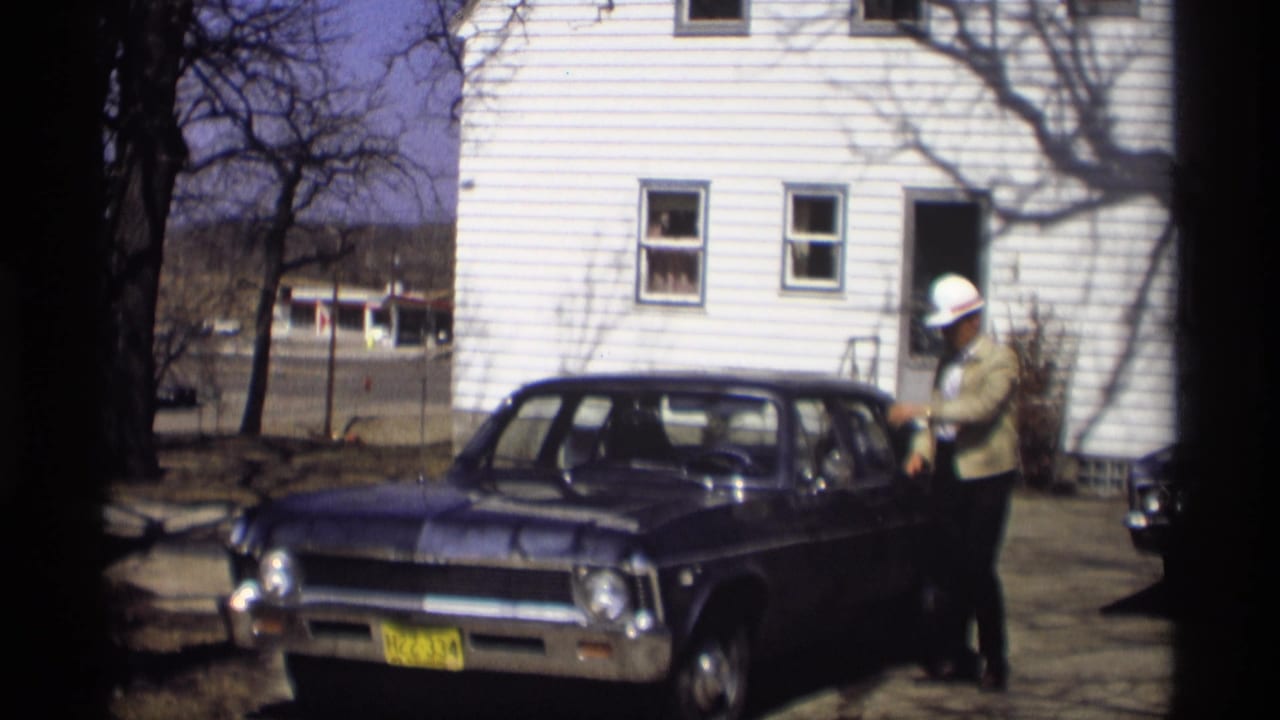 The man stands beside the blue sedan with a white hat on, framed by the white house and bare trees in Madison.