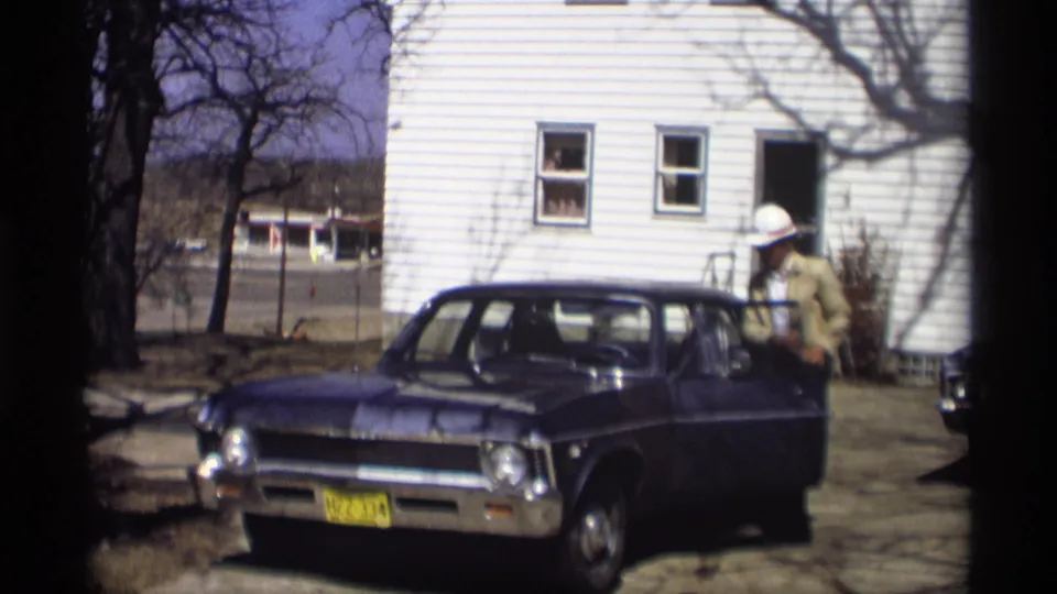 In the 1966 Madison home-movie scene, the man opens the driver's door of the blue sedan outside the house.