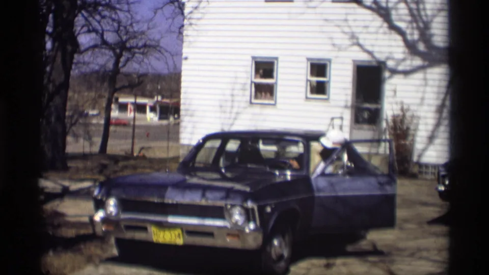 The man leans into the driver's seat of the blue sedan, with the white clapboard house and bare trees behind him.