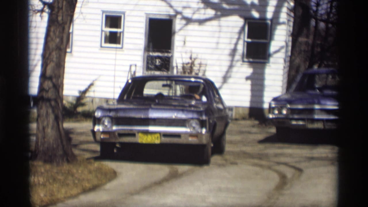 Blue sedan parked in a driveway before a white house with another car nearby.