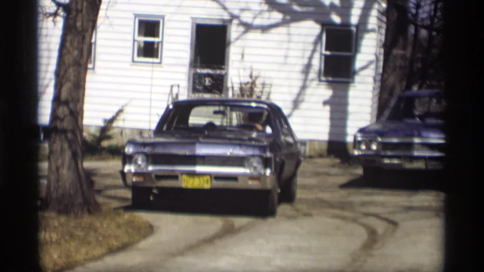 1966 Madison home-movie frame showing a blue Chevrolet Impala facing camera in a residential driveway beside a white-sided house.