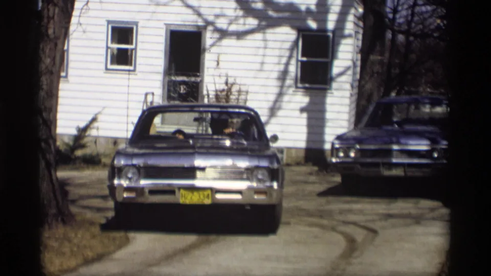 The blue Chevrolet Impala rolls forward on the residential driveway, framed by trees and the white-sided house in Madison.