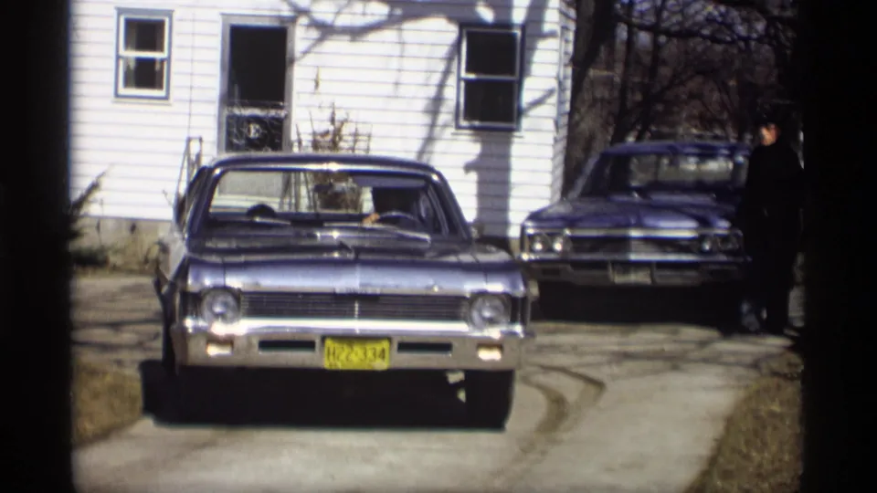 The blue Chevrolet Impala is closer in the driveway while a person appears beside the second car at the right edge of the frame.