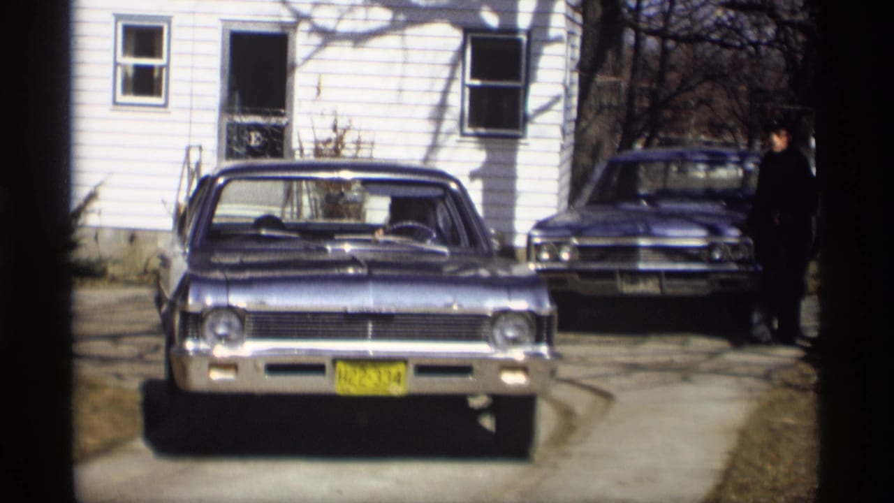 Blue sedan facing the camera in a driveway beside a white house, with another car and a man behind it.