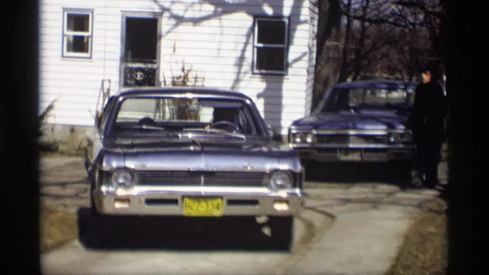 1966 Madison home-movie frame showing a blue sedan in a driveway near a white clapboard house, with a second car and man in back.