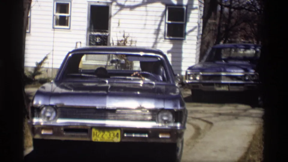 A blue mid-1960s sedan approaches the camera in a Madison driveway, with a white house and another parked car behind it.