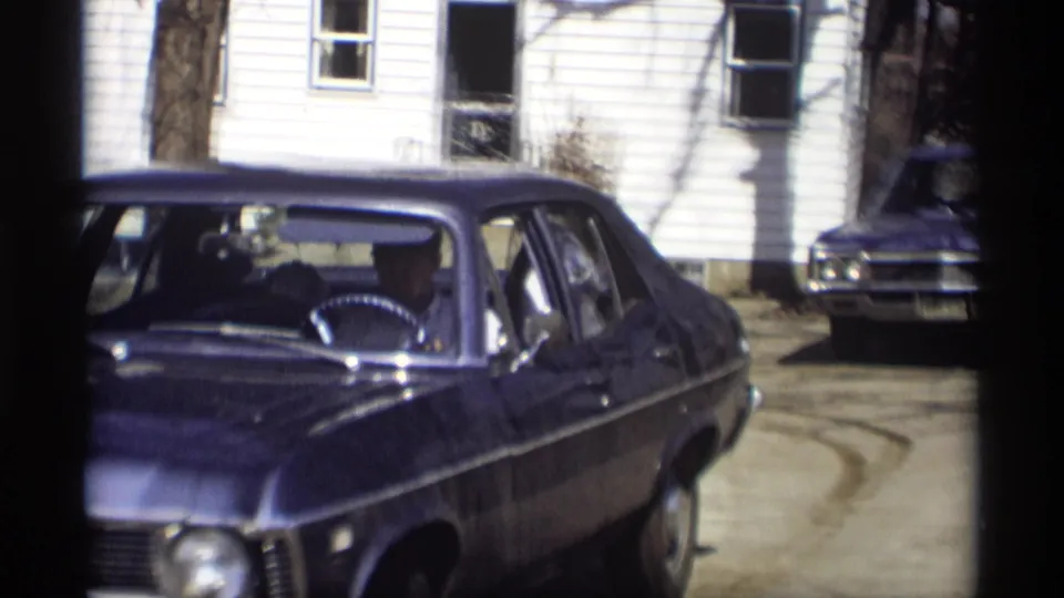 The blue sedan passes close in side view during a 1966 Madison driveway departure, with another car parked near the house.