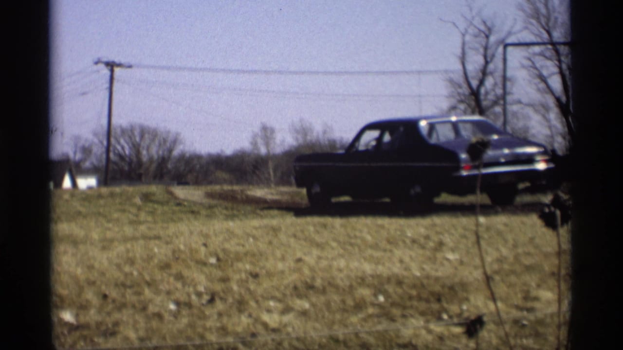 Dark sedan moving across a dry grassy field with fence strands, bare trees, and utility poles behind it.