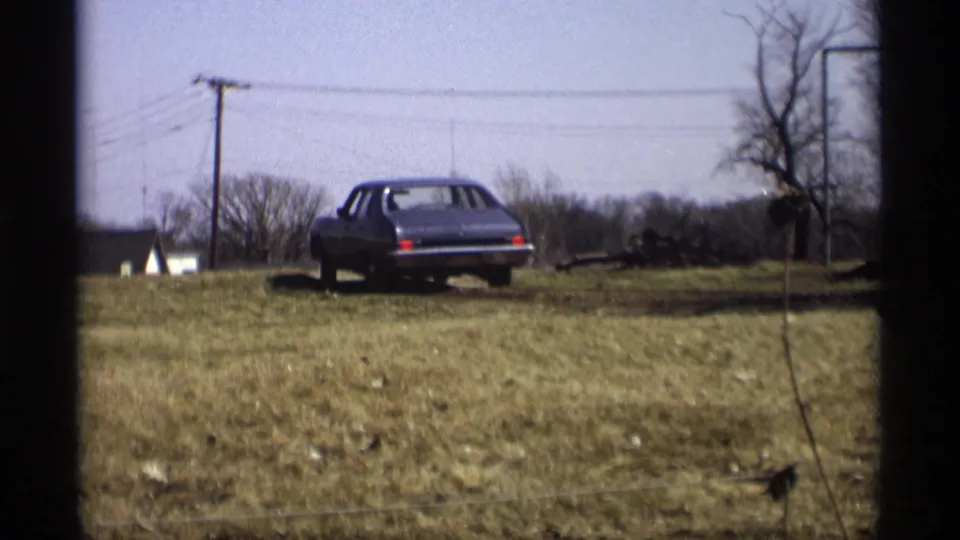 Rear of the sedan as it moves away across open grass, framed by leafless trees, utility wires, and the soft grain of 8mm film.