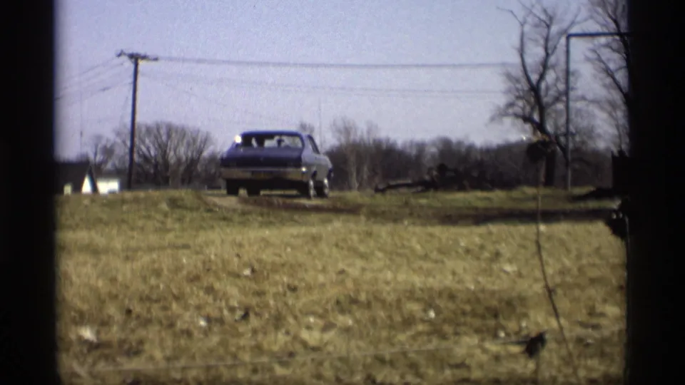 The sedan travels away through dry grass, with a small white building or roofline, utility pole, bare trees, and fence strands visible.
