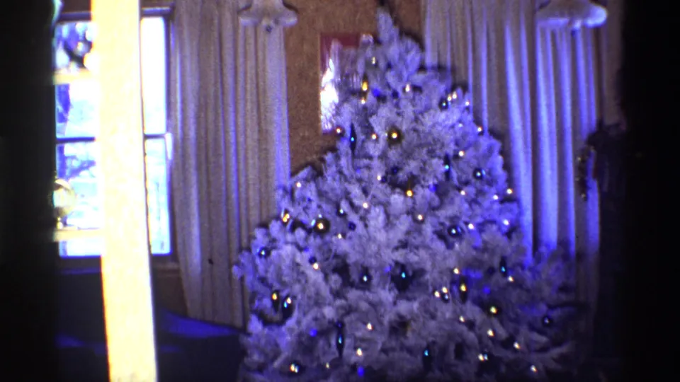 1968 Chicago home-movie frame showing a white Christmas tree with blue and gold ornaments in a softly lit interior.