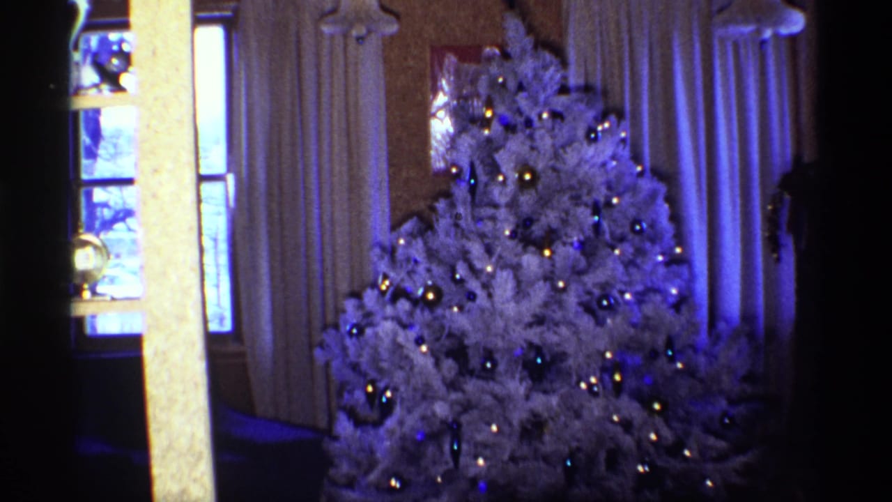 White Christmas tree with ornaments beside curtains and a window in a dim living room.