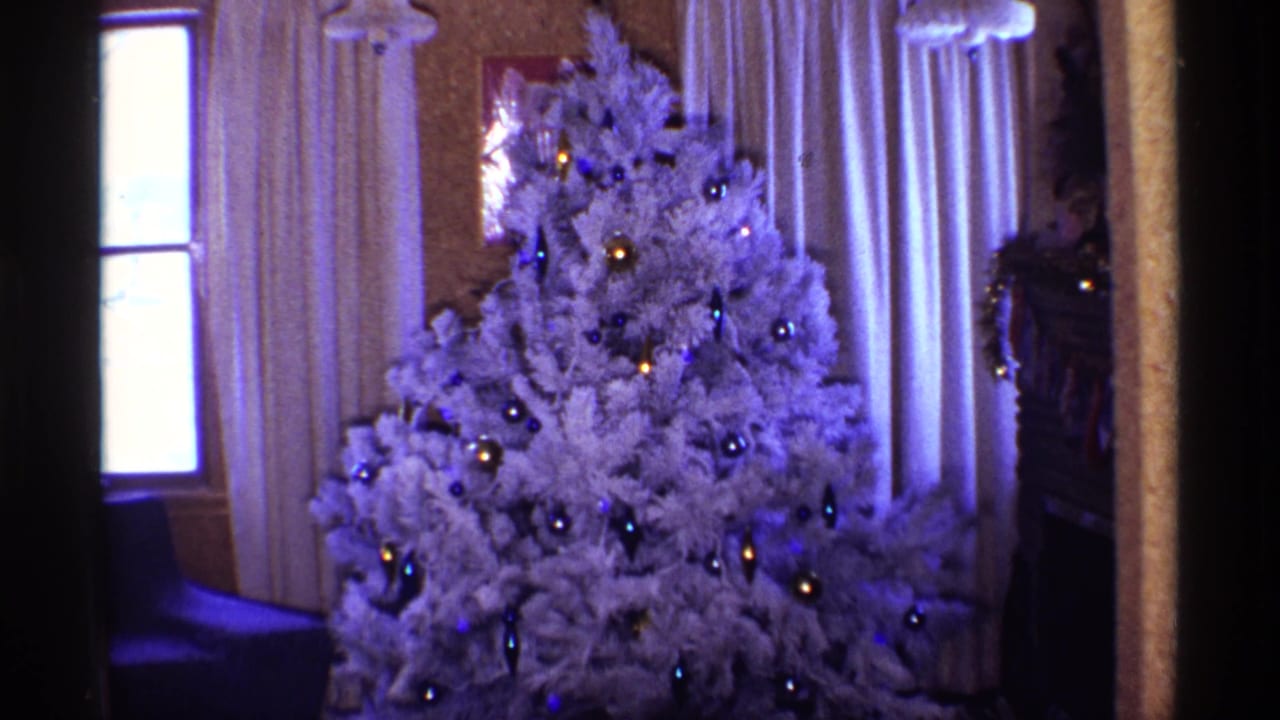 Flocked white Christmas tree with ornaments beside cream curtains and a window.