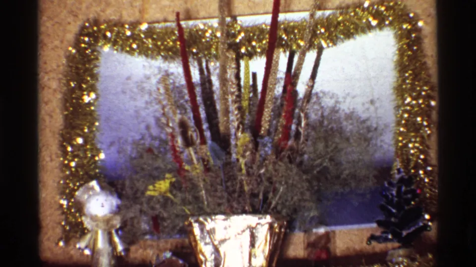 1968 Chicago home-movie frame of the mantel display, with gold garland framing the background and a foil vase holding tall decorative sticks.