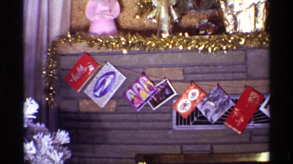 1968 Chicago home-movie frame showing the fireplace and mantel with gold garland, hanging holiday cards, small figures, foil vase, and a white artificial tree nearby.