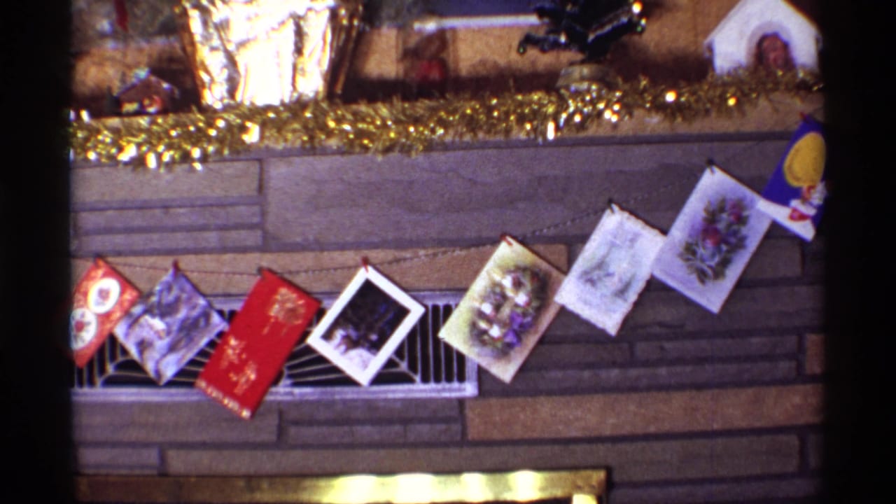 1968 Chicago frame showing a diagonal string of Christmas cards across a stone fireplace, with metallic garland and holiday objects on the mantel.
