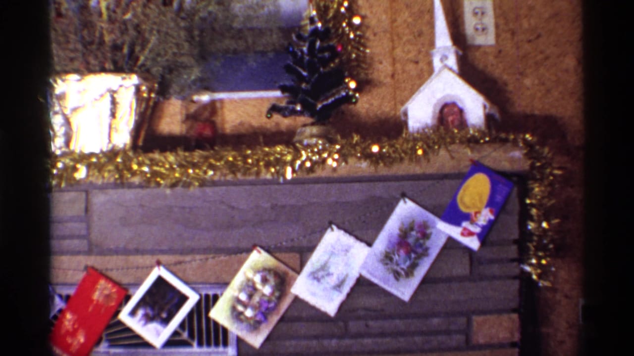 1968 Chicago home interior frame showing a decorated Christmas fireplace mantel with card garland, gold tinsel, foil planter, small tree, and church model.