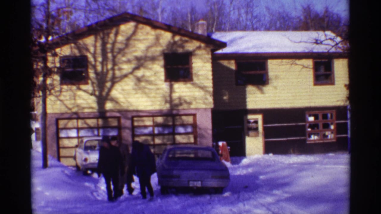 The men remain near the cars on the snowy driveway, framed by a yellow-sided house, garage doors, and bare winter trees.