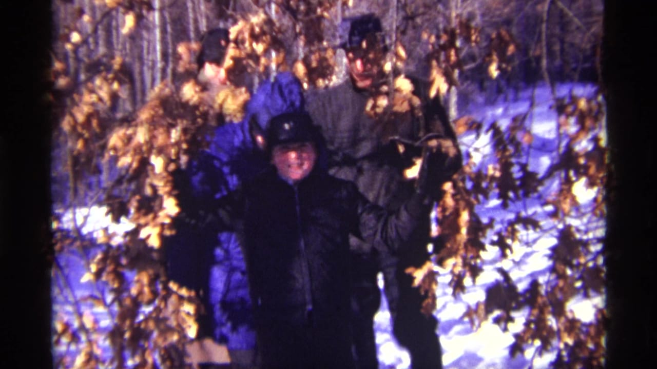 Bundled people walking through snowy woods under bare trees with dry leaves.