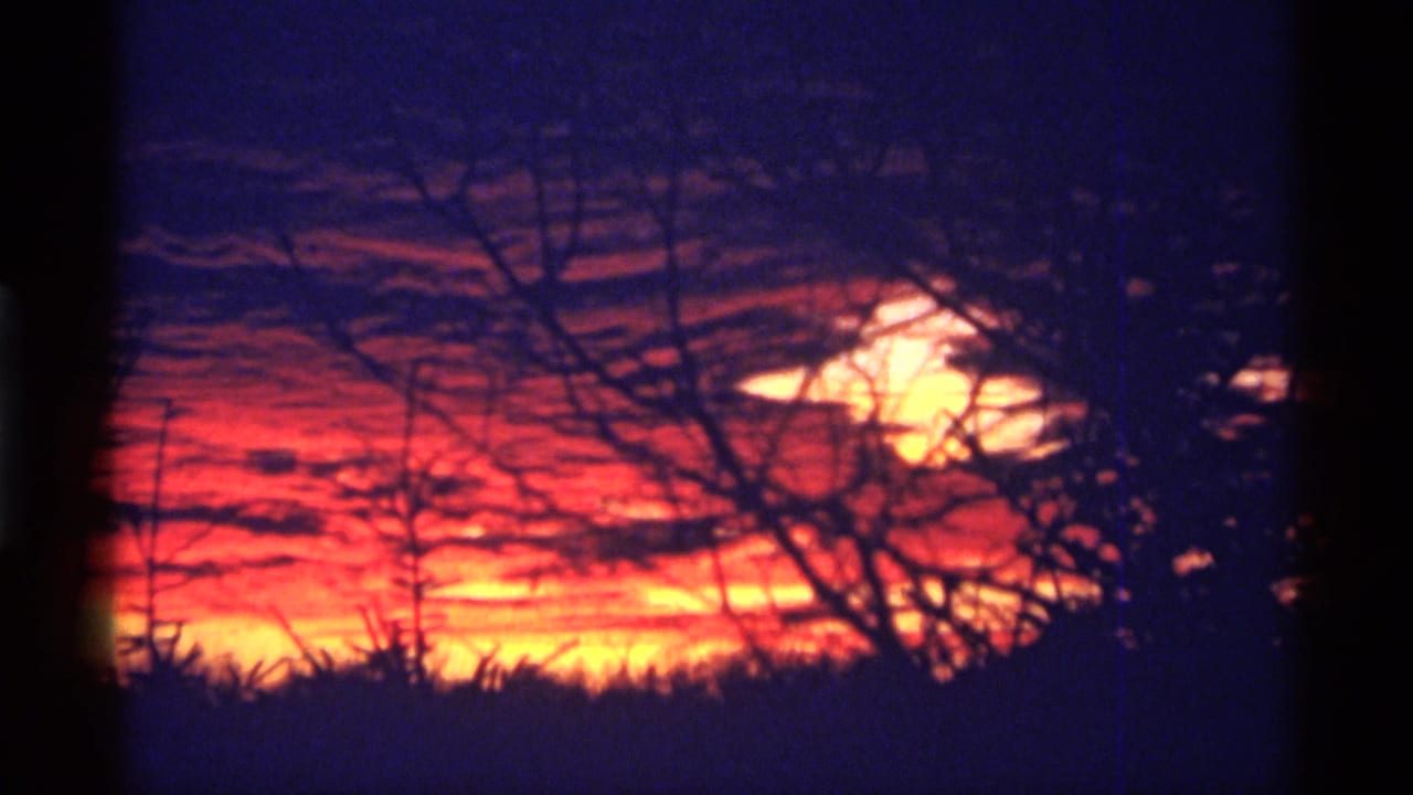 1968 8mm home-movie frame from Chicago metadata showing orange-red sunset clouds, purple-blue sky, and silhouetted winter branches.