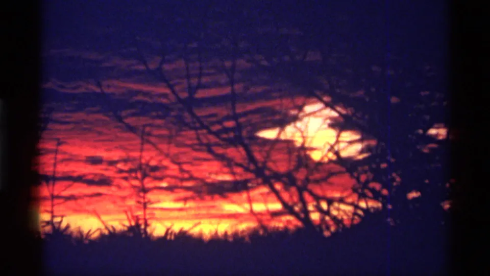 1968 8mm home-movie frame from Chicago metadata showing orange-red sunset clouds, purple-blue sky, and silhouetted winter branches.