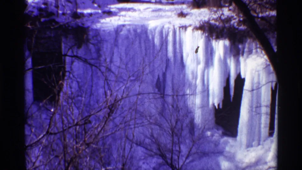 Blue-toned 8mm frame of a frozen waterfall in winter, with long ice columns, bare branches, and shadowed tree trunks.