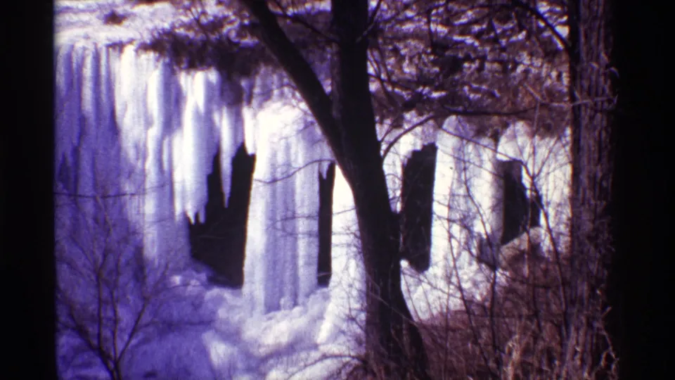 Chicago-area winter home-movie frame showing thick frozen falls, dark foreground trees, tangled branches, and snow-covered ice below.