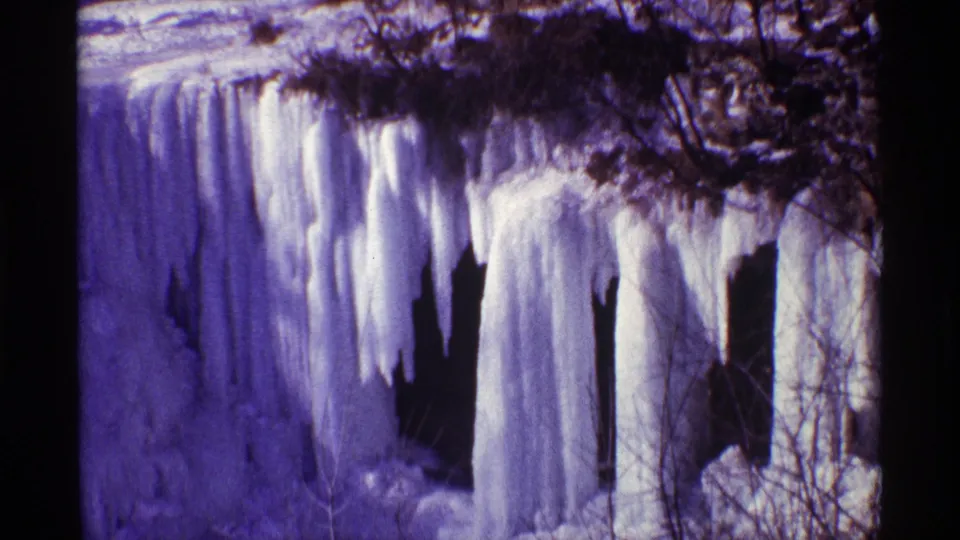 1968 8mm frame showing dense icicles, frozen runoff, bare brush, and shadowed openings in the rock or bank.