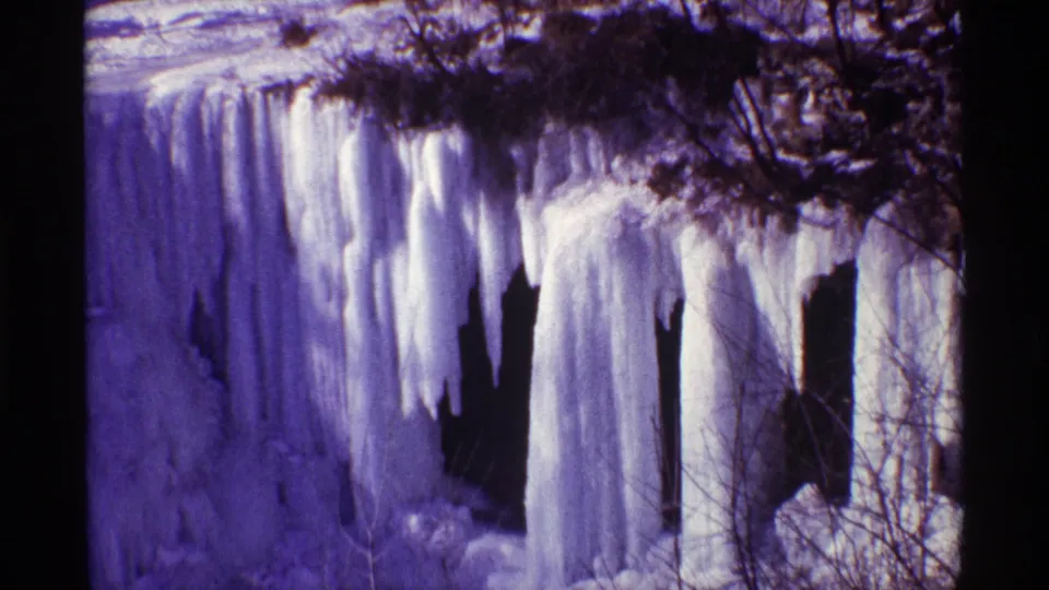 Chicago winter home-movie frame showing tall icicles and frozen water framed by a dark trunk and leafless branches.