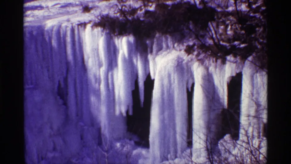 1968 8mm frame near Chicago showing thick icicles and frozen water hanging from a dark rocky cliff with winter branches.
