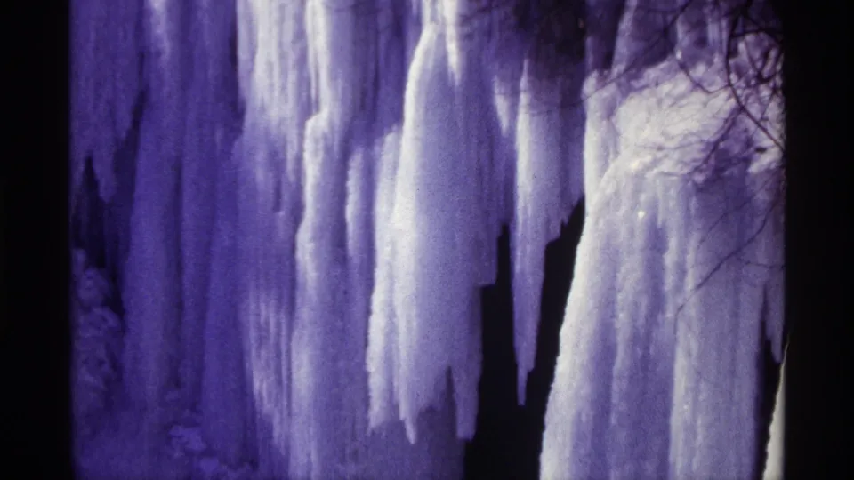 1968 8mm frame showing a tall wall of frozen water and icicles beside dark rock, with leafless branches at the edge.