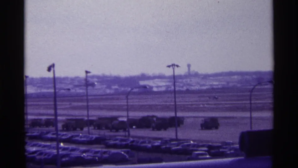 1968 Chicago airport frame showing parked cars, light poles, and a small distant airplane on or near the tarmac.