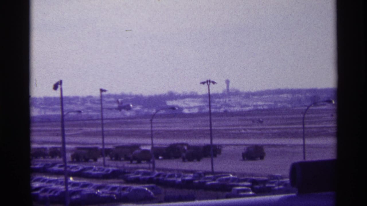 1968 Chicago airport view with a commercial airplane beginning takeoff beyond parked cars and service vehicles.