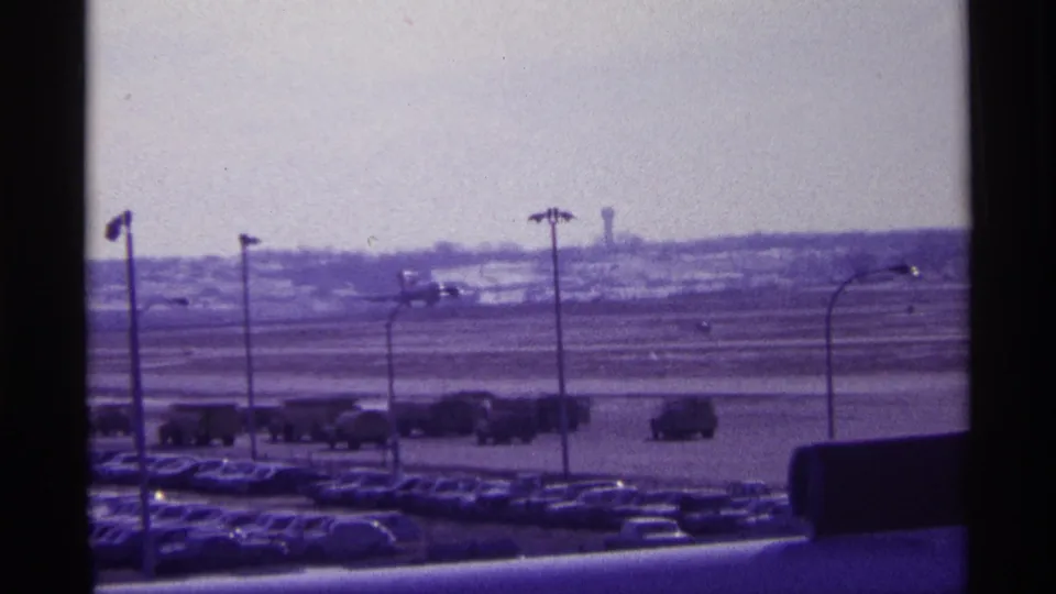 Distant 1968 Chicago airport runway scene with a plane climbing over parked cars, light poles, and tarmac vehicles.