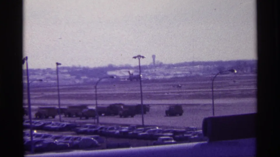 Soft 8mm view of a Chicago airport runway in 1968, with a distant plane, parking lot, service vehicles, and light poles.