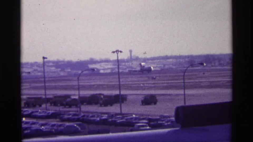 1968 Chicago airport footage showing a plane at distance beyond the runway, with parked cars and tarmac vehicles below.