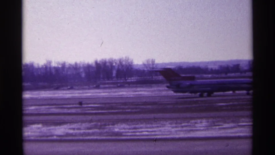 1968 Chicago airport frame showing a jetliner continuing along a winter tarmac with bare trees beyond the runway.