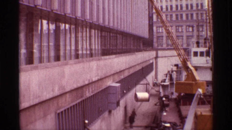 A yellow dock crane lifts a cylindrical load along a Chicago waterfront building, with workers and a cargo vessel below.