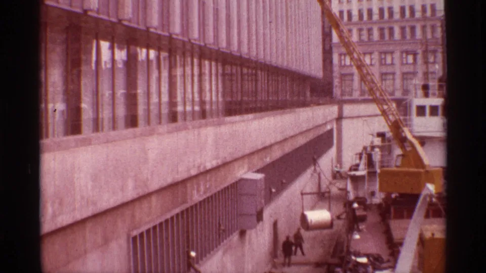 Workers move along the dock below a modern waterfront building while the crane positions cargo near the vessel.