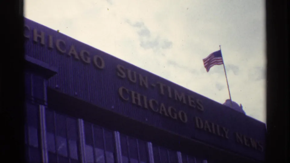 1968 Chicago 8mm frame showing newspaper mastheads on a building facade with an American flag against the sky.