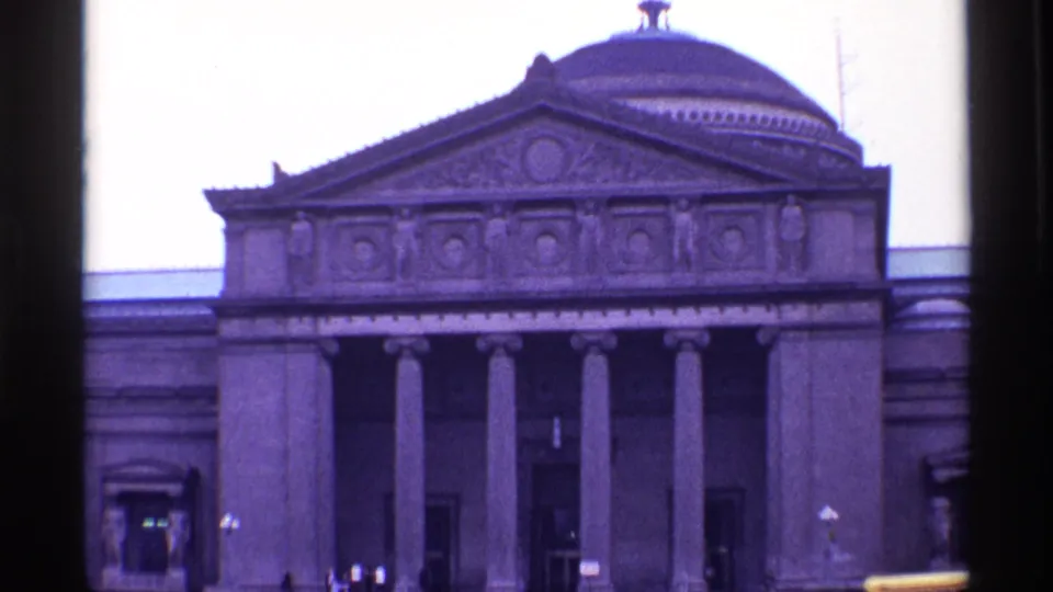 Home-movie frame of a Chicago classical-style building with a dome, columned entrance, and a yellow vehicle passing at street level.